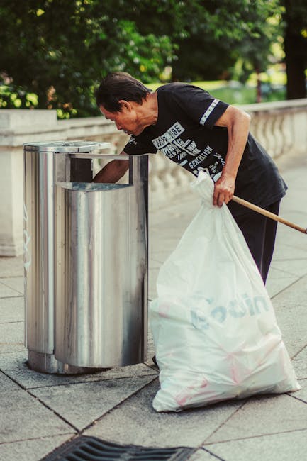 A middle-aged man with dark hair wearing a black t-shirt with white lettering is seen disposing of waste into a tall, cylindrical, stainless steel rubbish bin on a paved sidewalk. The bin has a polished, reflective surface and features a rectangular slot on the front for waste disposal. The man is holding a large white plastic bag filled with rubbish in his left hand, while he leans forward to insert trash through the opening. His right hand holds a wooden-handled litter picker, resting on the bag. The background includes a low concrete barrier, lush green trees, and a grassy area, suggesting an outdoor urban setting. Natural daylight illuminates the scene, highlighting the textures of the metal bin and pavement. This image illustrates the process of on-site waste collection by individuals, reflecting private or independent rubbish disposal practices often associated with alternative waste handling or rubbish removal services like those offered by House Clearance Soho.