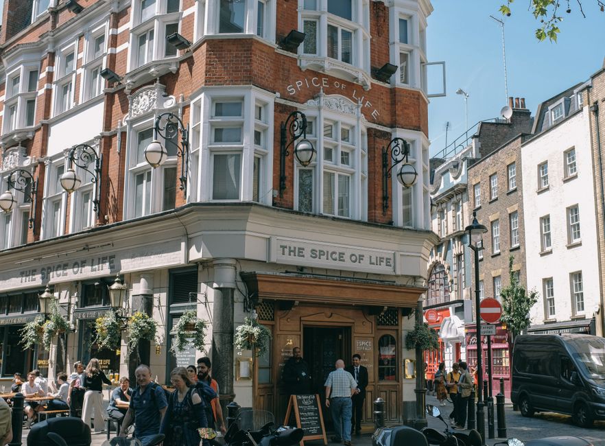 The exterior of a multi-story building in an urban setting, featuring red brickwork and white decorative window frames. The ground floor has a restaurant named 'The Spice of Life,' with a beige facade and black signage, including outdoor seating with several tables occupied by people dining. Hanging flower baskets and outdoor lamps are mounted on the building's façade, contributing to a lively, welcoming atmosphere. Pedestrians walk along the sidewalk, some engaged in conversation, while a few stand near the entrance of the restaurant. To the right, a dark delivery van is parked on the street, and a red telephone booth is visible in the background among other buildings, including a mix of brick and white façades. The scene is well-lit with natural daylight, capturing the bustling character of a city street with a focus on the lively public space in front of the restaurant. This setting reflects a typical urban environment where independent eateries and pedestrian activity coexist, aligning with local waste management needs associated with busy hospitality establishments.