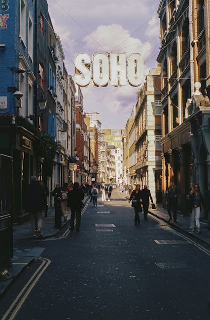 The image depicts a lively urban street scene in Soho, with a wide pedestrianized walkway bordered by multi-story buildings featuring varied architectural styles, including brick and plaster facades in shades of beige, brown, and dark grey. The street is populated by several people walking, some alone, others in groups, dressed in casual and semi-formal attire, suggesting a typical busy day in the area. In the background, a large, illuminated sign spelling 'SOHO' hangs overhead, suspended between buildings, casting a soft glow against the partly cloudy sky with hints of blue. The ground surface of the street appears dark and textured, possibly asphalt, with subtle markings and curbside edges visible. To the right, a building with ornate detailing and large windows indicates a retail or commercial establishment. The scene overall conveys a vibrant, mixed-use urban area that may involve the routine collection and disposal of waste and rubbish, often coordinated through private waste management services such as House Clearance Soho, which operates in this part of London.