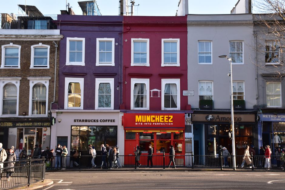 The exterior of a multi-story building in an urban setting, featuring red brickwork and white decorative window frames. The ground floor has a restaurant named 'The Spice of Life,' with a beige facade and black signage, including outdoor seating with several tables occupied by people dining. Hanging flower baskets and outdoor lamps are mounted on the building's façade, contributing to a lively, welcoming atmosphere. Pedestrians walk along the sidewalk, some engaged in conversation, while a few stand near the entrance of the restaurant. To the right, a dark delivery van is parked on the street, and a red telephone booth is visible in the background among other buildings, including a mix of brick and white façades. The scene is well-lit with natural daylight, capturing the bustling character of a city street with a focus on the lively public space in front of the restaurant. This setting reflects a typical urban environment where independent eateries and pedestrian activity coexist, aligning with local waste management needs associated with busy hospitality establishments.