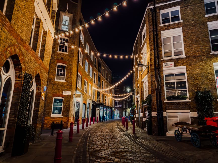 A nighttime view of a narrow cobbled street lined with multi-storey brick buildings featuring large sash windows with white frames, some with window boxes, and decorative brickwork. String lights are suspended overhead, casting a warm glow over the scene. The street is partially enclosed by black and red bollards along the edges of the pavement, with a few small trees and potted plants near the buildings. On the right side, there is a black vintage-style street lamp mounted on the wall, and a red wheelbarrow is resting against the wall. The scene exudes a quiet, atmospheric urban ambiance typical of a London alleyway, with the scene slightly illuminated by ambient lighting from nearby shop windows and the decorative string lights, capturing a sense of a well-maintained, historic area potentially suitable for private rubbish removal or on-site clearance activities by companies like House Clearance Soho.