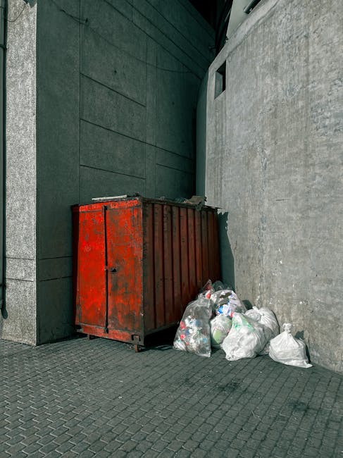 A weathered red metal skip positioned against a dark green wall and a plain grey concrete wall in an outdoor urban setting. The skip has visible rust and chipped paint, with its surface showing scratches and signs of use. Nearby, several white plastic rubbish bags filled with waste are piled on the grey textured pavement, some leaning against the base of the skip. The environment appears clean but utilitarian, with a small, dark opening or window visible in the grey wall behind the skip and no other objects or signage present. The scene suggests an area designated for private waste collection, typical of independent rubbish removal services like House Clearance Soho, set in a modern urban alley or backstreet space, optimized for discreet rubbish disposal in line with Westminster Council rules for Soho waste management.